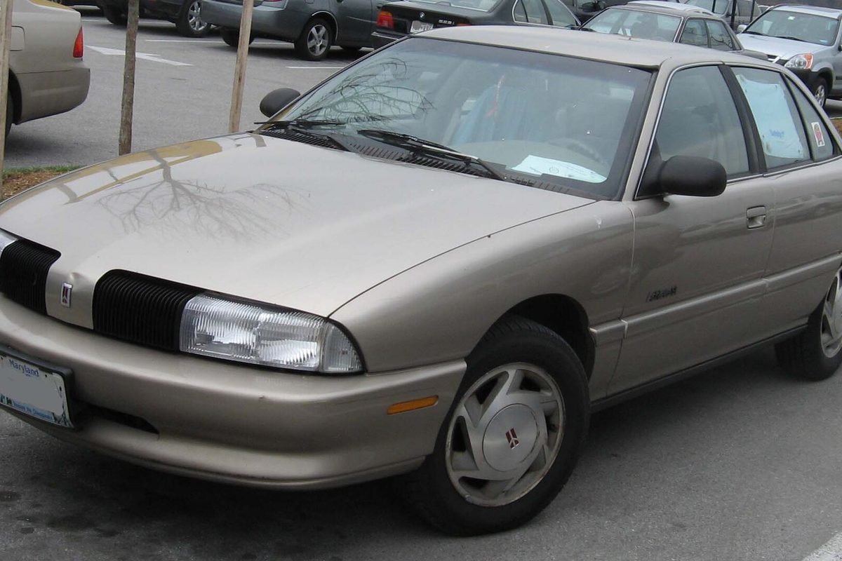 A tan Oldsmobile Achieva sedan with silver alloy wheels parked in a lot, a parking slip on the dashboard