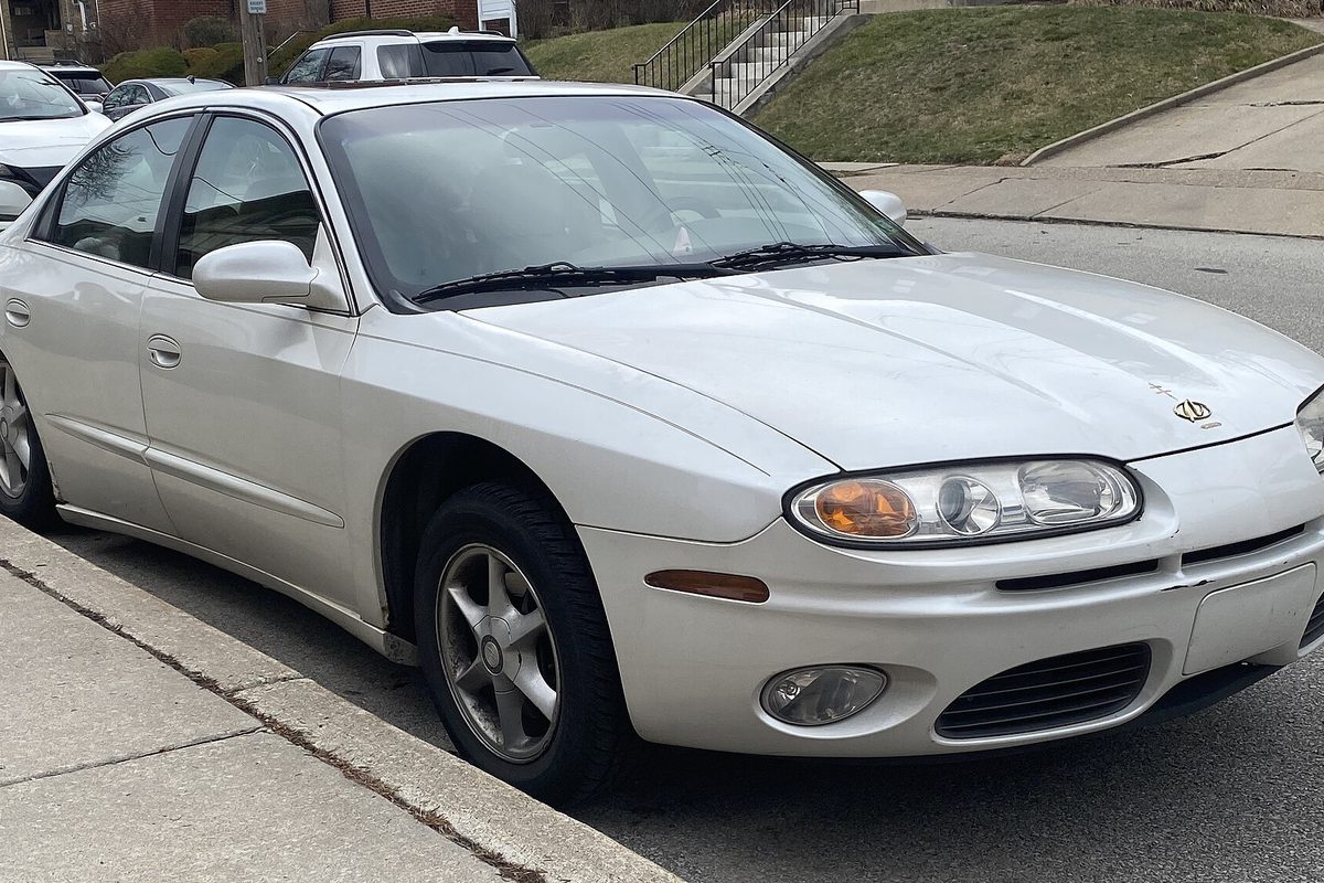A white 2001 Oldsmobile Aurora sedan parked at a curb on a residential street, seen from the front right
