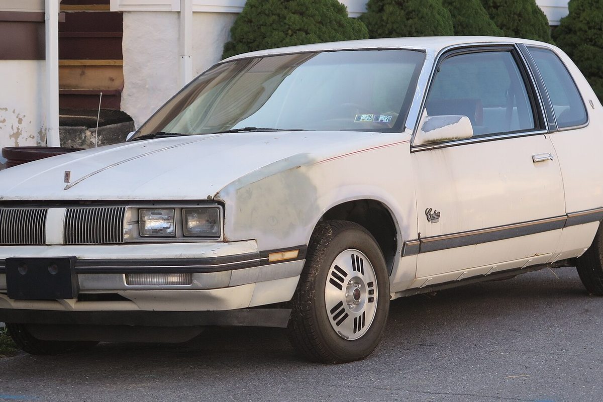 A white 1985 Oldsmobile Calais coupe with gray primer on the front fender, parked in a driveway beside shrubs