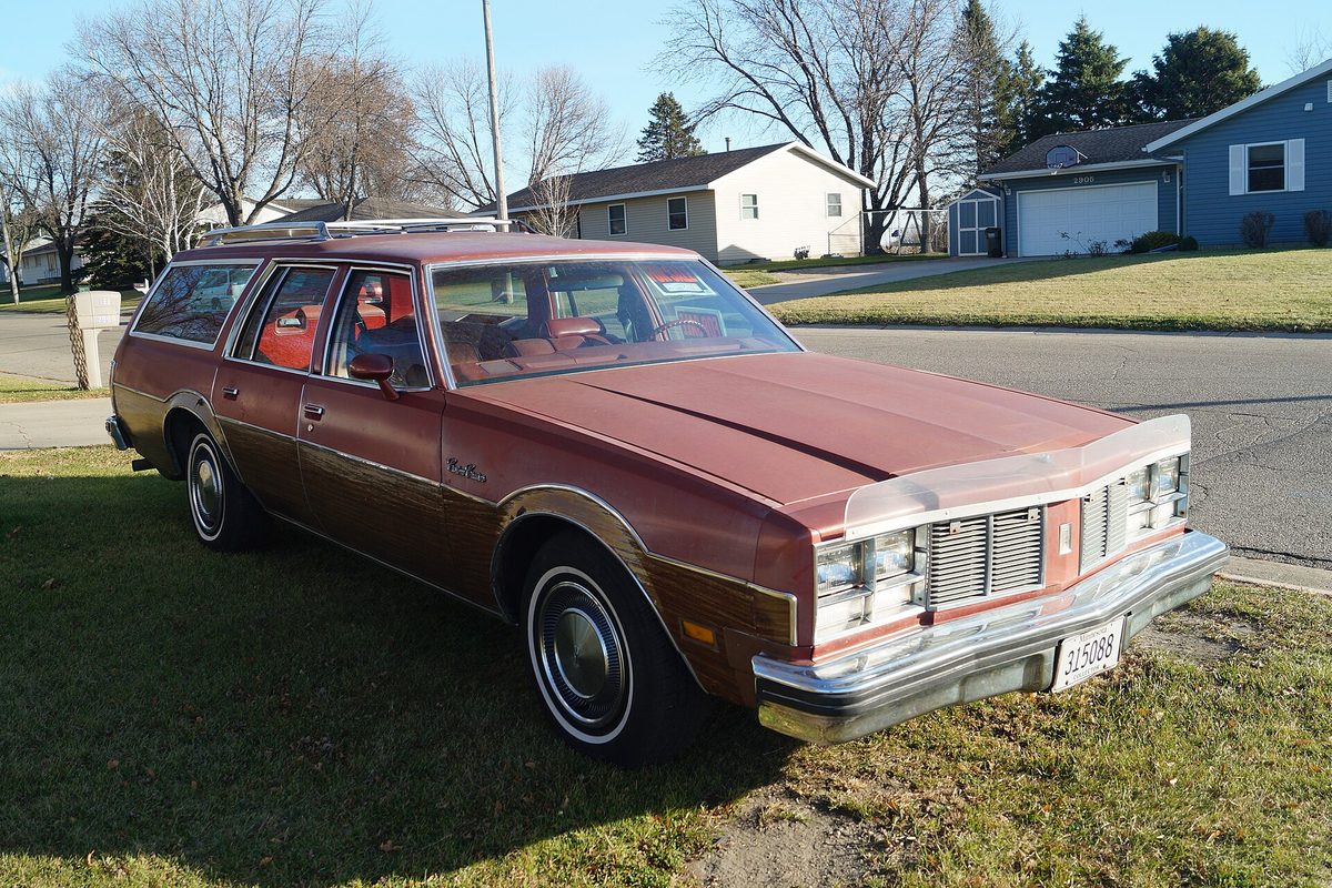 A maroon Oldsmobile Custom Cruiser station wagon with woodgrain side panels and a roof rack, parked on a lawn beside a quiet residential street