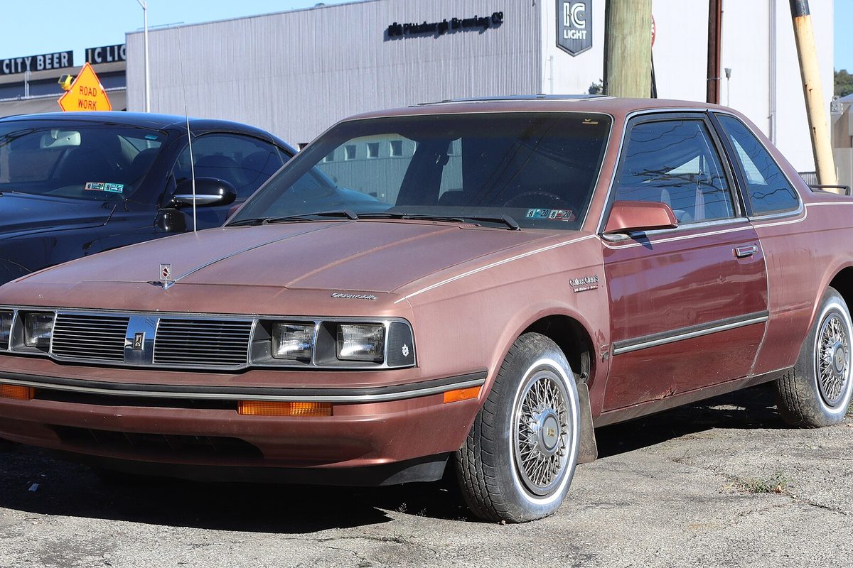 A light chestnut 1986 Oldsmobile Cutlass Ciera coupe with wire wheel covers and chrome trim, parked on a cracked lot