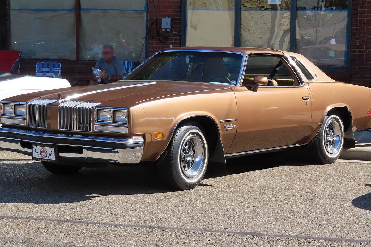 A saddle-brown 1976 Oldsmobile Cutlass Supreme Brougham coupe with chrome wheels and whitewall tires at a street car show