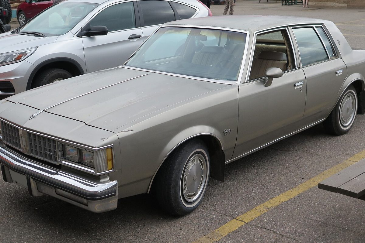 A silver-gray older-generation Oldsmobile Cutlass four-door sedan with a chrome grille and bumper, parked in a lot beside a newer car