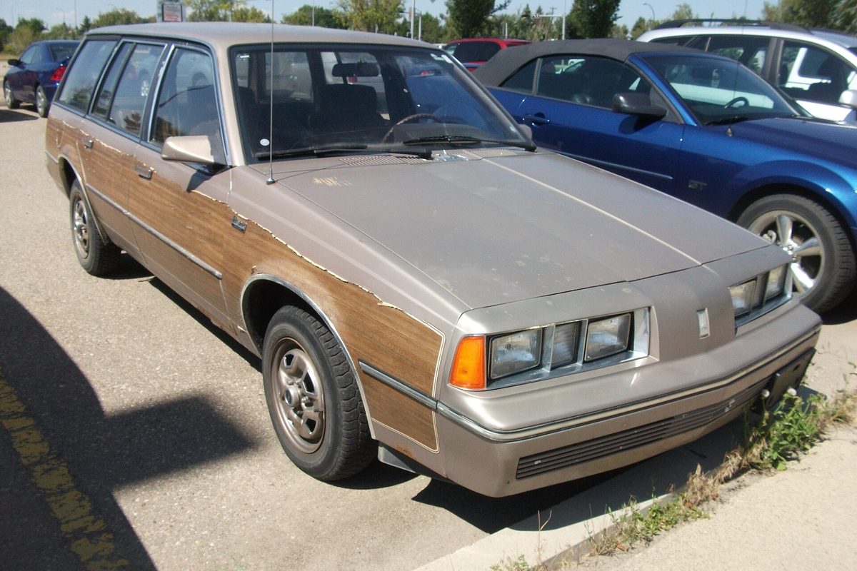 A tan Oldsmobile Firenza Cruiser station wagon with peeling woodgrain side panels, parked in a lot next to newer cars