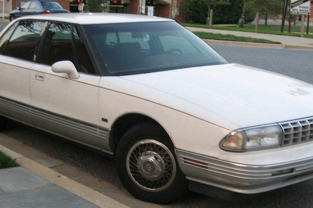 A white 1991-1996 Oldsmobile Ninety-Eight sedan with wire wheel covers and chrome trim, parked at a curb