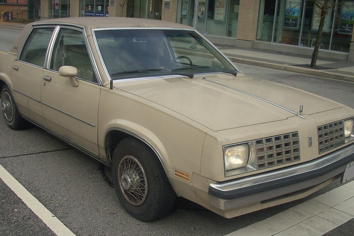 A beige third-generation Oldsmobile Omega four-door sedan with wire wheel covers, parked at a city curb