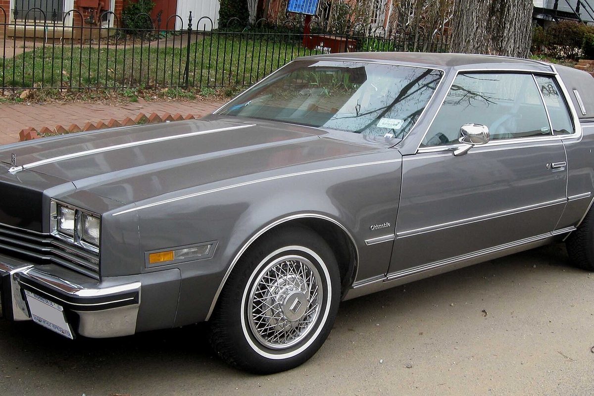 A gray 1979-1985 Oldsmobile Toronado coupe with a long hood, whitewall tires and wire wheel covers, parked by an iron fence on a city street