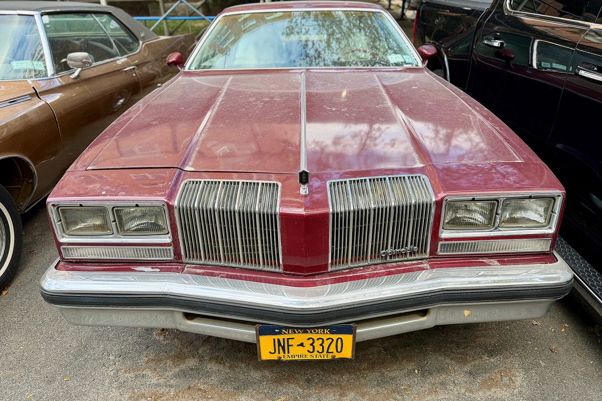 A maroon 1976 Oldsmobile Cutlass Supreme seen head-on in a parking lot, showing its split vertical-bar grille and stacked square headlights