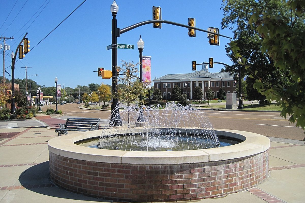 A round brick fountain at a downtown Olive Branch intersection, with the red brick City Hall and its white columns behind it under a clear blue sky