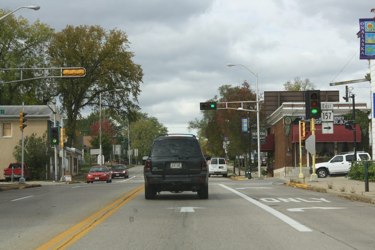 Traffic lights over a two-lane street in Onalaska where Highway 157 begins, with a dark SUV waiting at the signal and a brick storefront on the corner