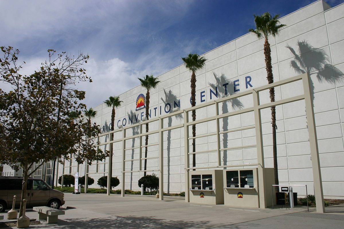 The white wall of the Ontario Convention Center with its name in large letters, tall palm trees and their shadows along the front, and a blue sky
