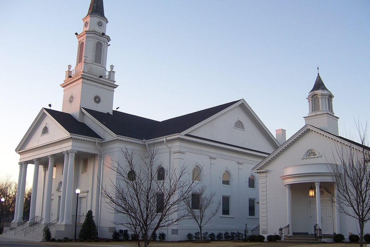 The white columned First Baptist Church of Opelika with its tall steeple and a smaller cupola, lit by early morning light under a clear sky