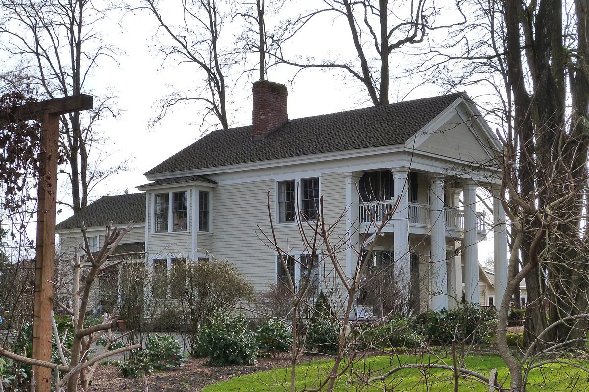 The white two-story Ainsworth House in Oregon City, with tall columns, a second-floor balcony and bare winter trees around it