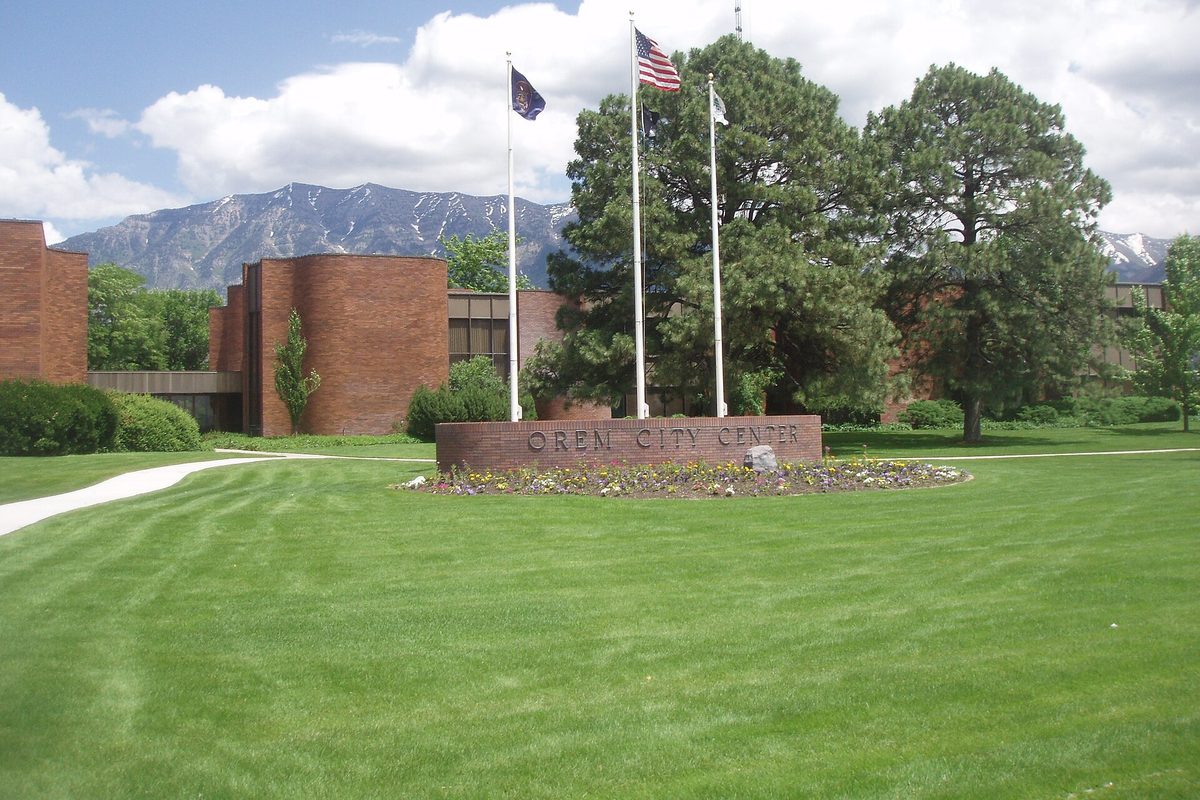 The Orem City Center sign in a flower bed on a wide green lawn, with flagpoles, pine trees, brick buildings and rocky mountains behind