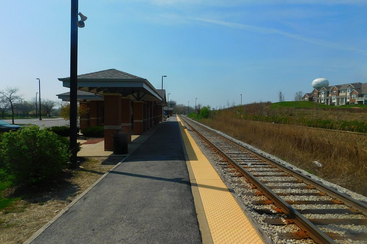 The empty platform and tracks at Orland Park's 153rd Street Metra station on a clear day, with a brick shelter and a water tower in the distance