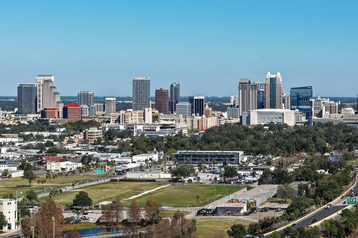 Downtown Orlando's high-rise skyline seen from the west on a clear day, with trees, a ball field and a highway in the foreground