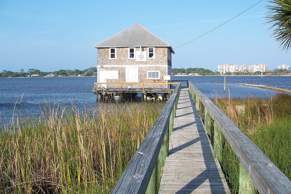 A weathered shingled boathouse on pilings over the water at the end of a wooden boardwalk through tall marsh grass, with high-rise buildings far across the water