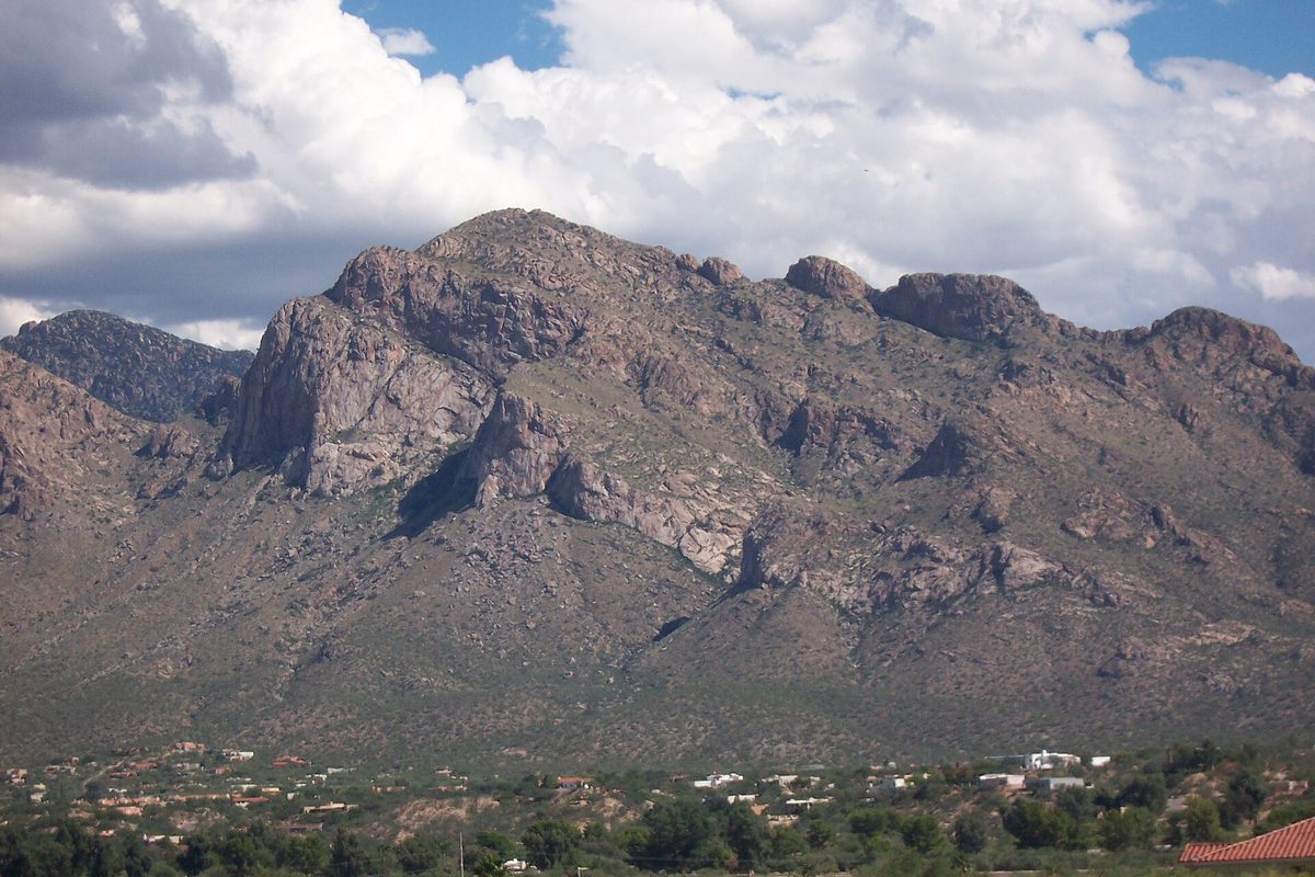 The rocky face of Pusch Ridge in the Santa Catalina Mountains under white clouds, with Oro Valley homes and trees along its base