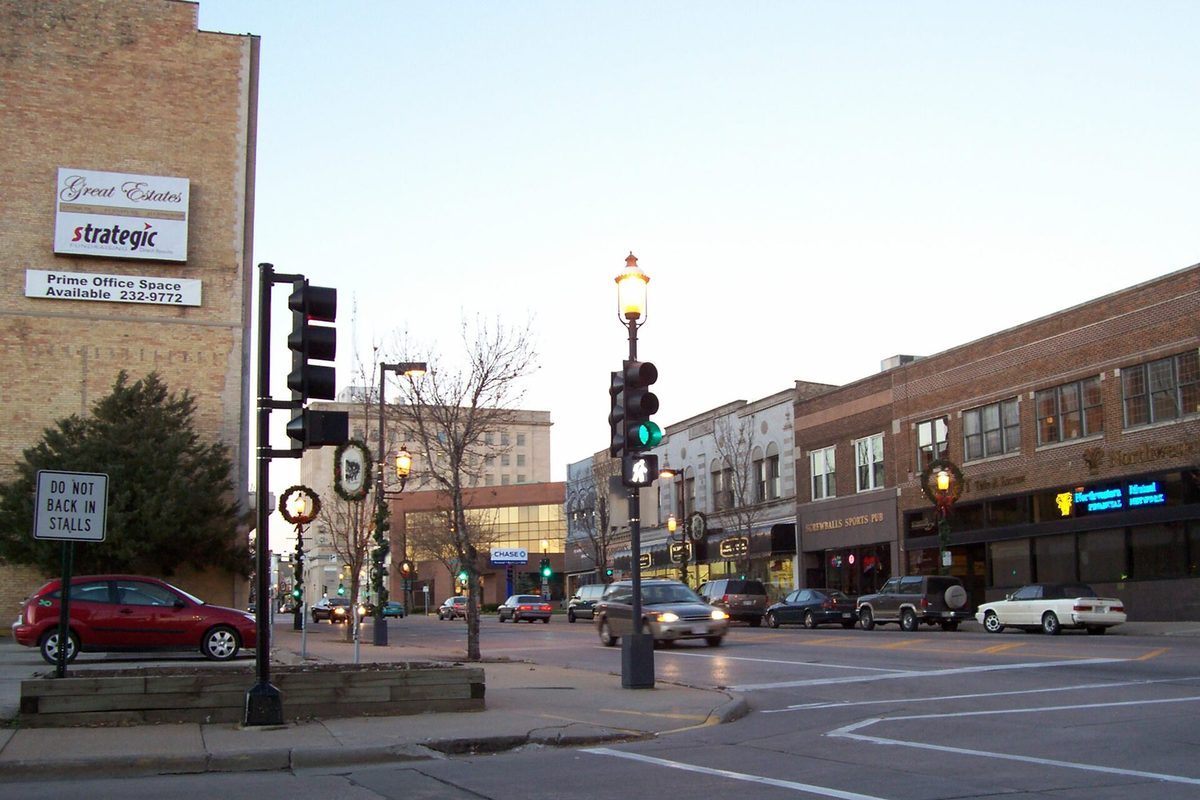Downtown Oshkosh at dusk, looking along Main Street past brick storefronts, traffic lights and holiday wreaths on the lamp posts, with a red car parked at the corner
