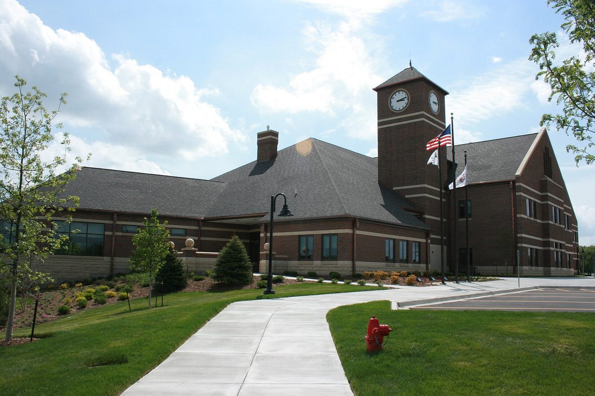 Oswego Village Hall, a brick building with a clock tower and flags, behind a curving sidewalk, green lawn and a red fire hydrant
