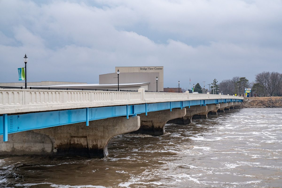 A white bridge with blue girders over the high, brown Des Moines River in Ottumwa, with the Bridge View Center behind it under gray clouds