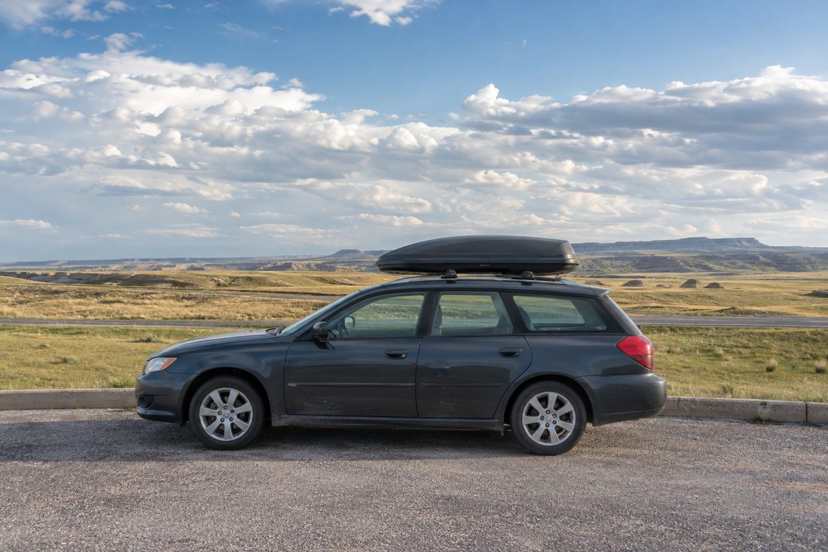 A dark station wagon with a roof cargo box parked beside a road with open grassland and distant buttes under a cloudy sky