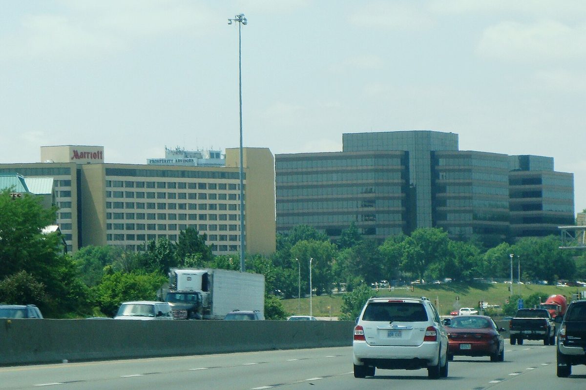 Traffic on I-435 in Overland Park, with an SUV and a box truck passing office towers and a hotel behind a concrete barrier and trees