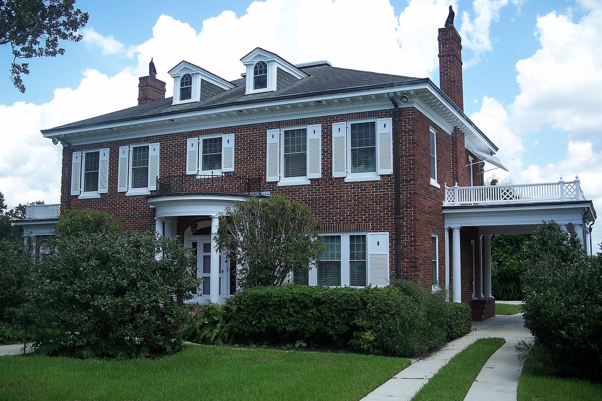 The two-story red brick Wheeler-Evans House in Oviedo, with white shutters, dormers, a side porch and trimmed shrubs on a green lawn