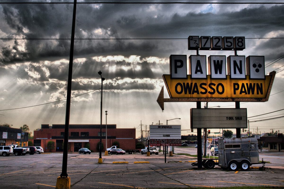 The tall Owasso Pawn sign against a dark, stormy sky with sunbeams breaking through the clouds, a utility trailer parked beneath it on an empty lot
