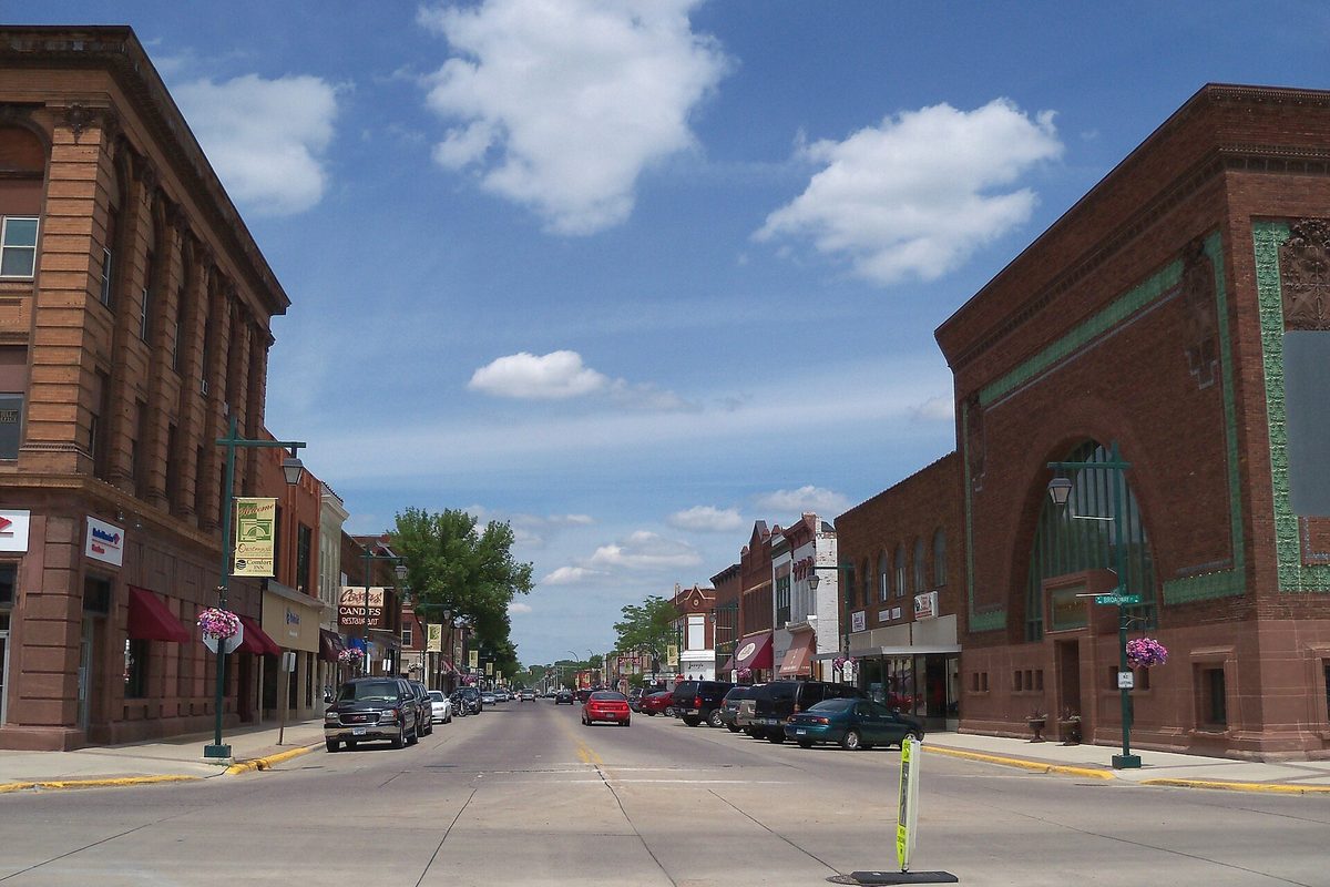 Downtown Owatonna's main street, lined with brick storefronts, flower baskets on lamp posts and parked cars, under a blue sky with white clouds