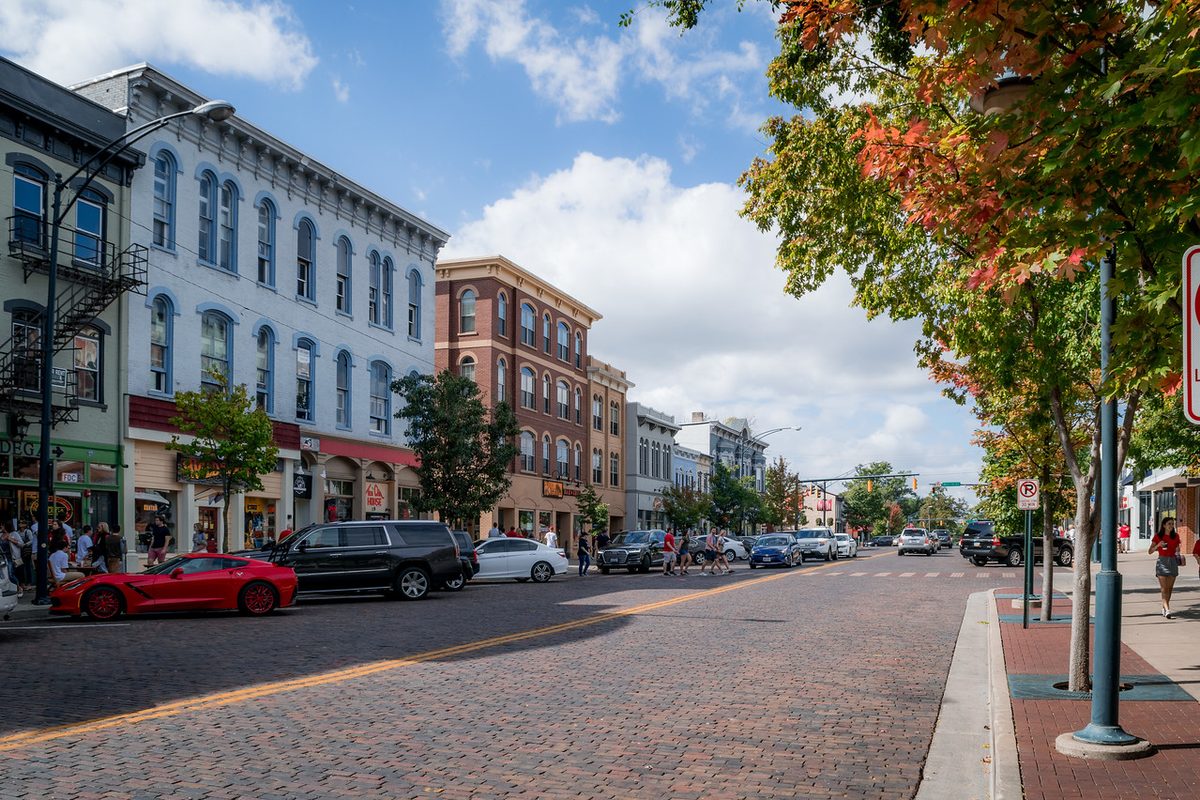 Uptown High Street in Oxford on a sunny fall day, with brick and painted storefronts, a red sports car at the curb and trees turning orange along the brick-paved street