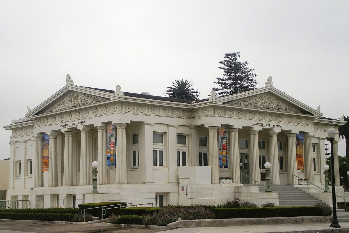 The white columned Carnegie Art Museum in Oxnard under a gray sky, with a palm and a tall pine rising behind its roofline