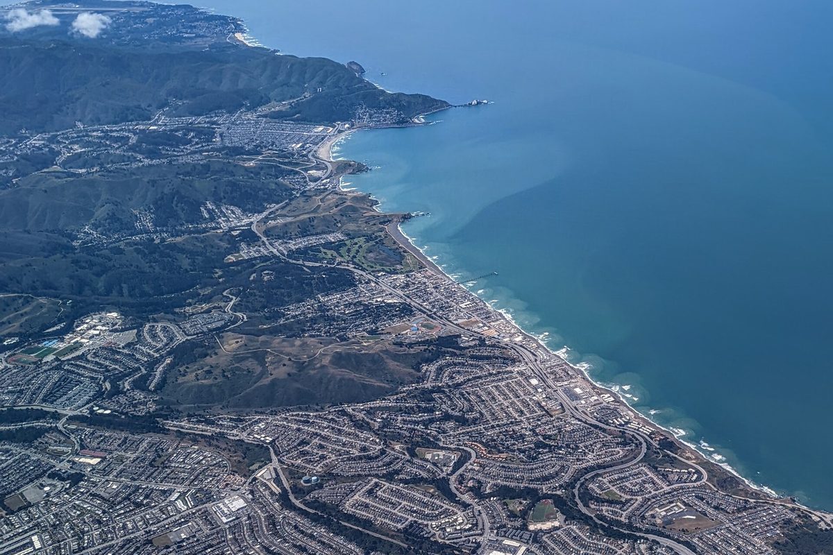Aerial view of Pacifica's neighborhoods and green ridges running down to the Pacific shoreline, with a pier jutting into blue water