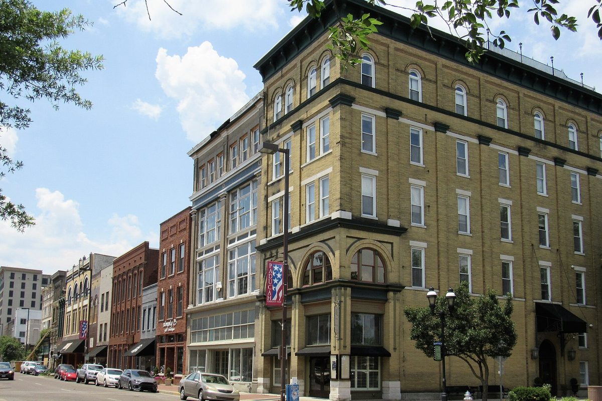 A tall tan brick building with arched windows on Broadway in downtown Paducah, older storefronts and parked cars along the street behind it