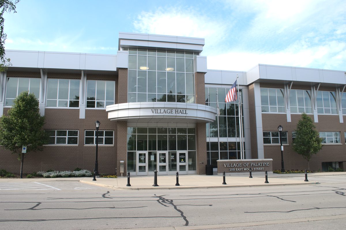 Palatine Village Hall, a two-story brick and glass building with a curved entrance canopy, an American flag and a sign reading Village of Palatine, 200 East Wood Street