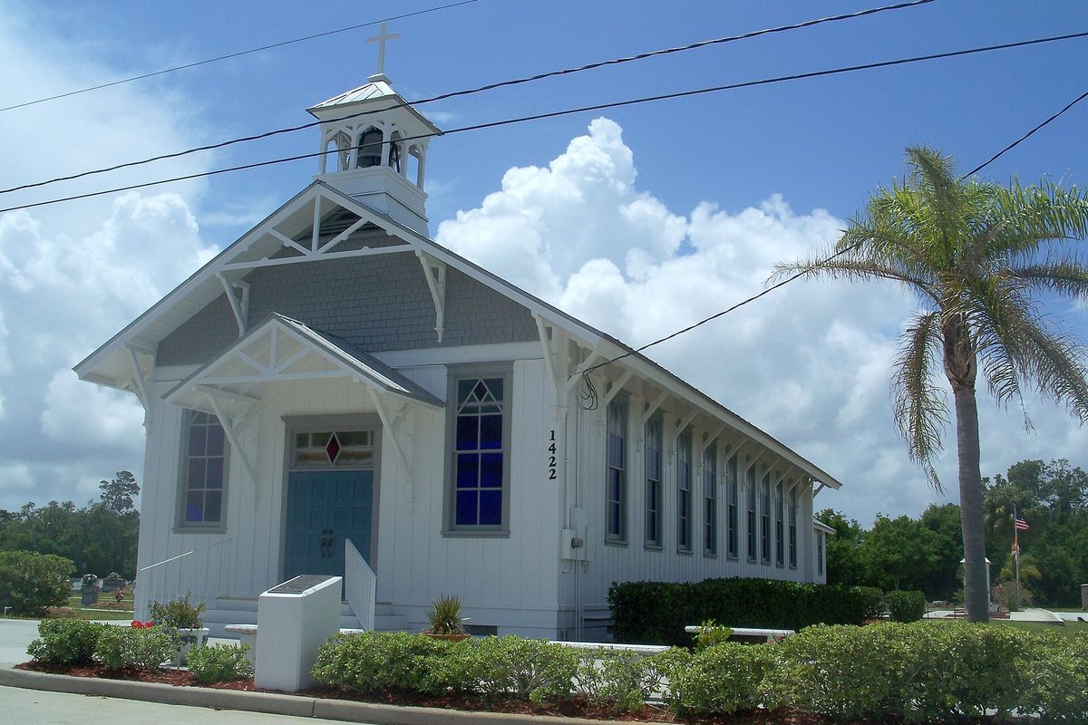 A white wooden church with a small bell tower, a blue door and tall narrow windows, next to a palm tree under a cloudy summer sky