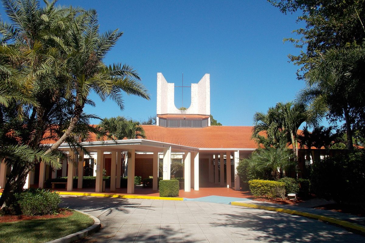 The Cathedral of St. Ignatius Loyola in Palm Beach Gardens, a white open bell tower above an orange tile roof and covered walkway, framed by palms