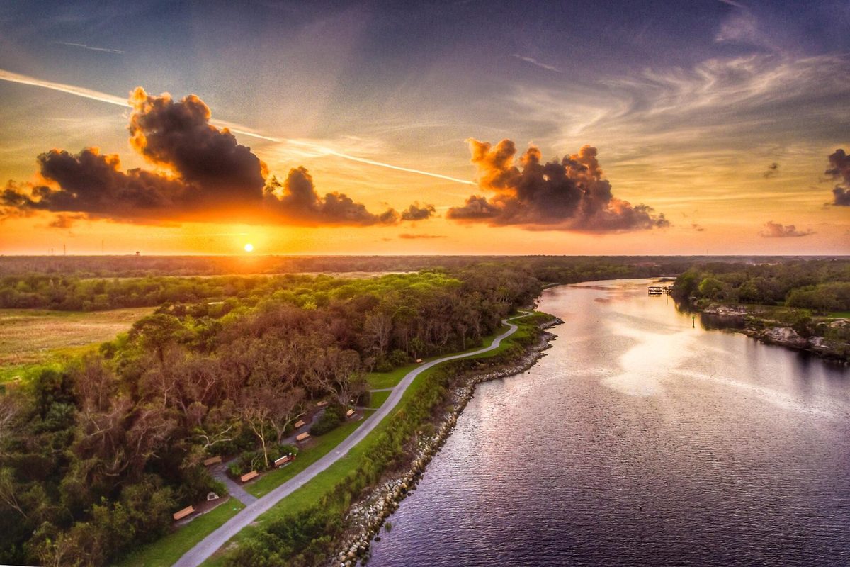Aerial view of a Palm Coast sunset over a calm waterway, with a paved path and picnic shelters running along a wooded shore