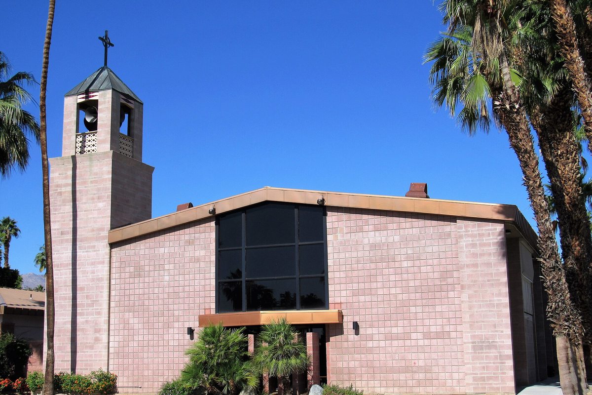 Sacred Heart Church in Palm Desert, a pink block building with a tall dark window and a bell tower topped by a cross, framed by palm trees under a deep blue sky