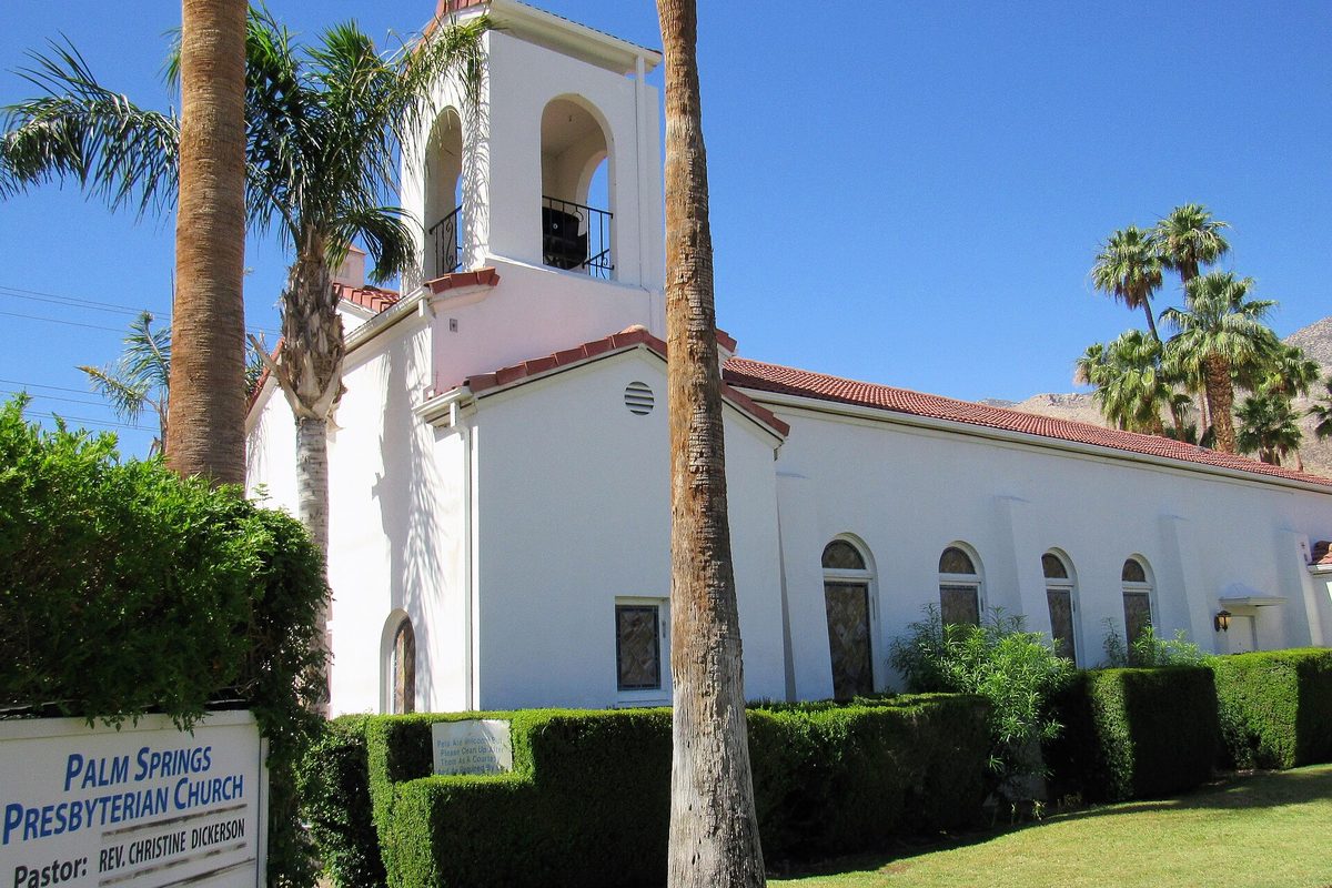Palm Springs Presbyterian Church, a white stucco building with a red tile roof and an open bell tower, with tall palms, trimmed hedges and desert mountains behind it
