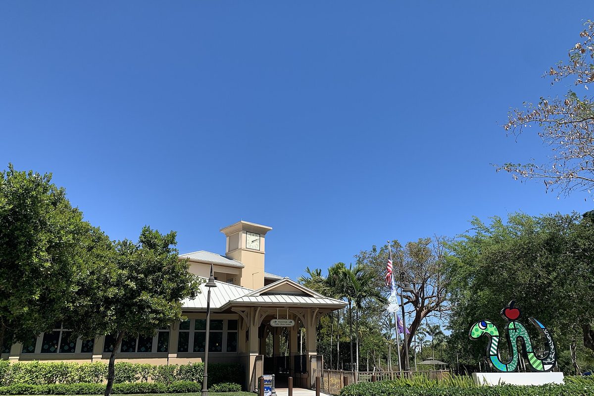 The Palmetto Bay Library, a tan building with a clock tower and metal roof, with a green snake sculpture holding a red apple in front under a clear blue sky