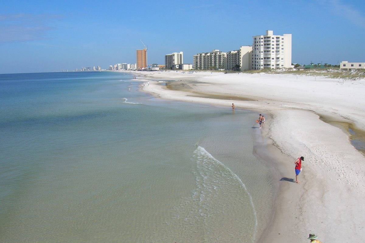 White sand beach and clear green Gulf water at Panama City Beach, with high-rise condo towers along the shore and a few people at the water's edge