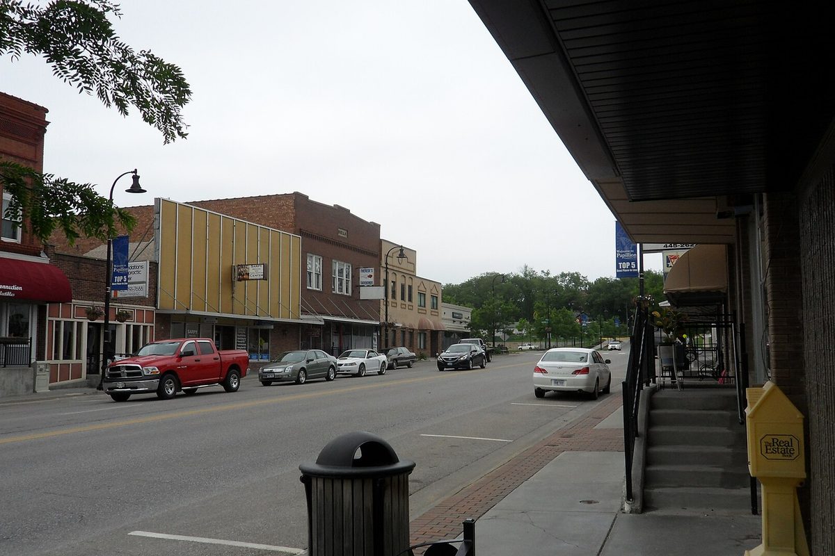 Washington Avenue in downtown Papillion, with brick and yellow-fronted storefronts, a red pickup and other cars parked at the curb on a cloudy day