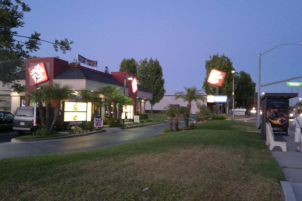 A fast-food restaurant with lit signs, palm trees and a lawn beside a bus shelter on a Paramount boulevard at dusk