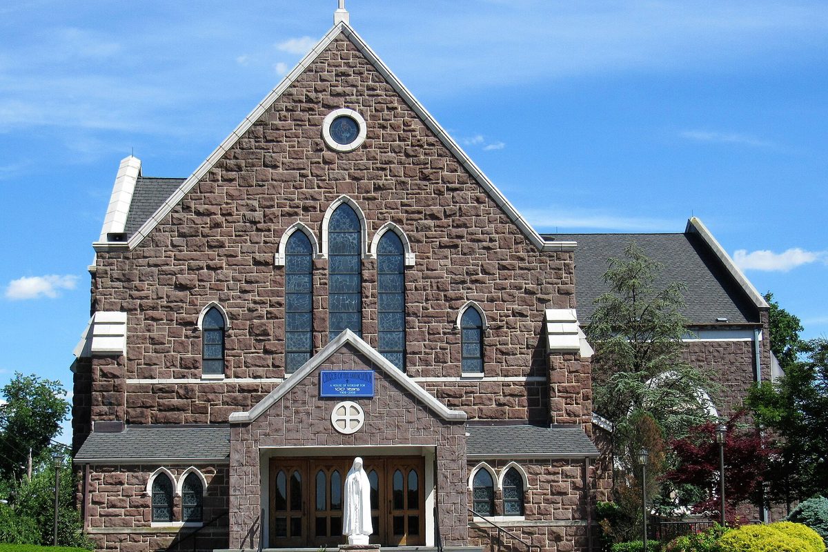The Church of the Annunciation in Paramus, a brown stone Gothic church with tall pointed stained-glass windows and a white statue at the entrance, under a blue sky