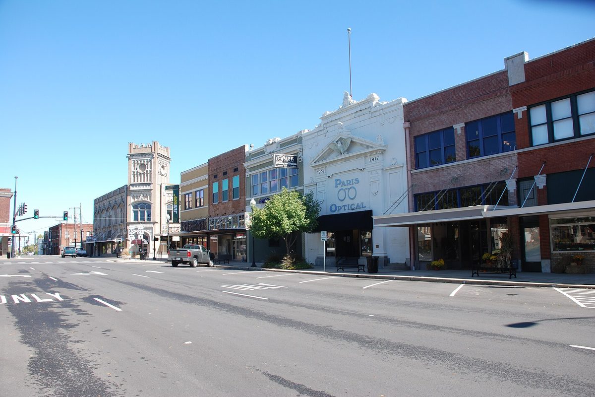 Main Street at the Plaza in Paris, Texas: two-story brick and white storefronts including Paris Optical, a pickup truck parked at the curb, an ornate tower down the block