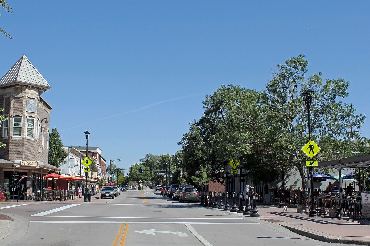 Mainstreet in Parker on a sunny day, with a brick corner building topped by a pointed turret, pedestrian crossing signs and patio umbrellas under shade trees