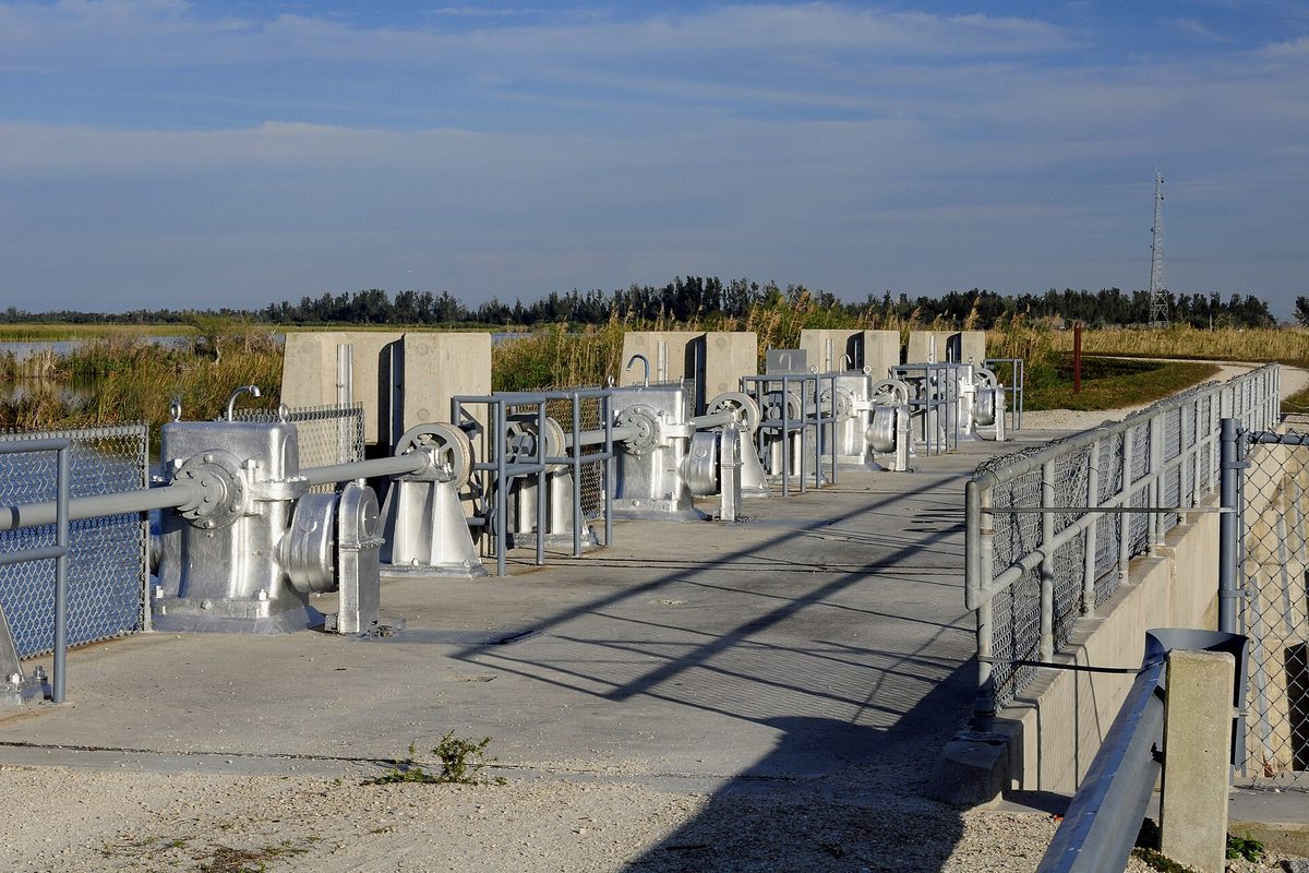 A row of silver gate-lift machines on a concrete water control structure beside a marsh, with a chain-link rail and a blue sky