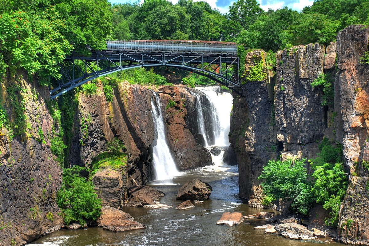 The Great Falls of the Passaic River in Paterson, water pouring over dark rock cliffs beneath a steel footbridge, with green trees above