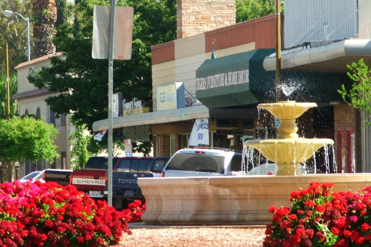 A tiered fountain splashing in a Patterson roundabout, ringed by red flowers, with storefronts and parked pickup trucks behind it